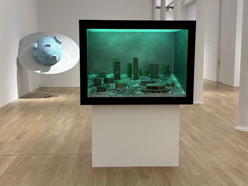 2 sculptures in an art gallery: one is a planetary sphere attached to a wall, the other is a green diorama of a city